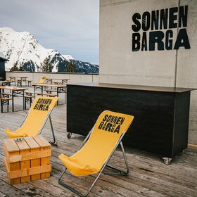 Der Eingang zur Sonnenbarga mit gelben Liegestühlen und Bierbänken auf der Terrasse. | © Silvretta Montafon - Vanessa Strauch