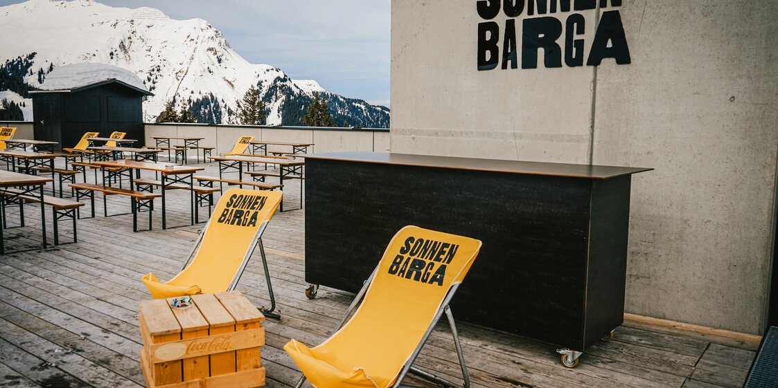 Der Eingang zur Sonnenbarga mit gelben Liegestühlen und Bierbänken auf der Terrasse. | © Silvretta Montafon - Vanessa Strauch