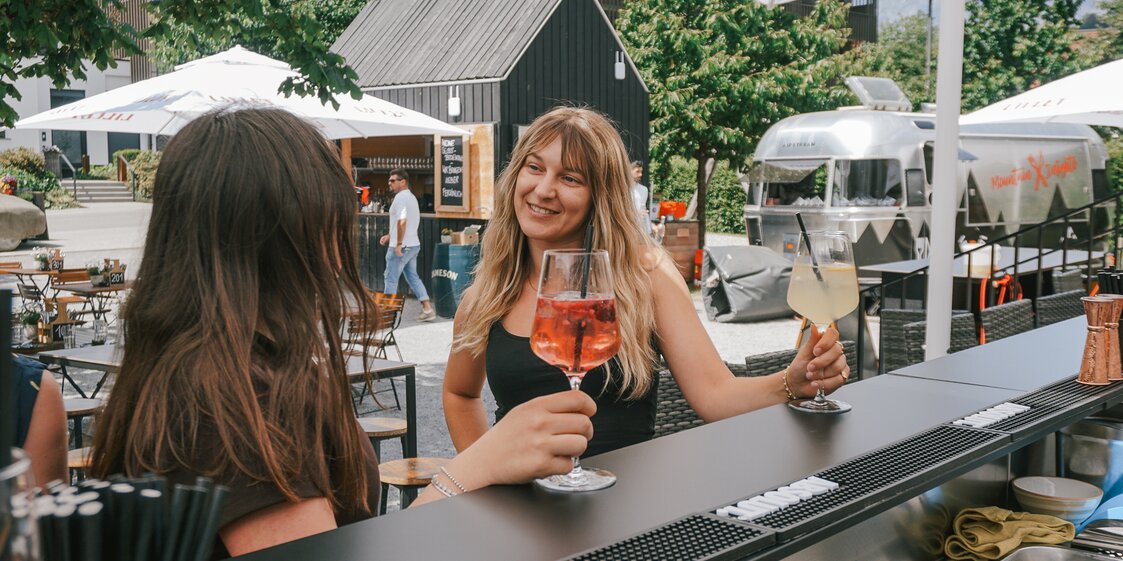 Zwei Frauen sitzen an einer Aussenbar in einem Gastgarten und trinken ein erfrischendes Sommergetränk aus einem Weinglas. | © Silvretta Montafon - Vanessa Strauch