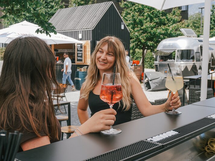 Zwei Frauen sitzen an einer Aussenbar in einem Gastgarten und trinken ein erfrischendes Sommergetränk aus einem Weinglas. | © Silvretta Montafon - Vanessa Strauch