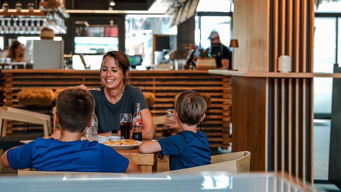 Eine Glückliche Familie sitz im Innenbereich der Markthalle und isst Pommes. | © Silvretta Montafon - Vanessa Strauch