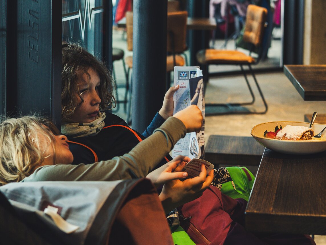 Zwei Jungen sitzen im Restaurant und schauen ein Prospekt an, auf dem Tisch steht ein Apfelstrudel mit Vanillesoße. | © Silvretta Montafon - Roadtyping