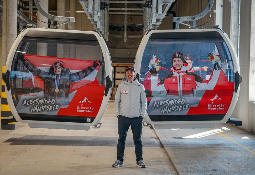 Alessandro Hämmerle steht neben zwei gebrandeten Gondeln mit seinen Olympiasiegen | © Silvretta Montafon - Vanessa Strauch