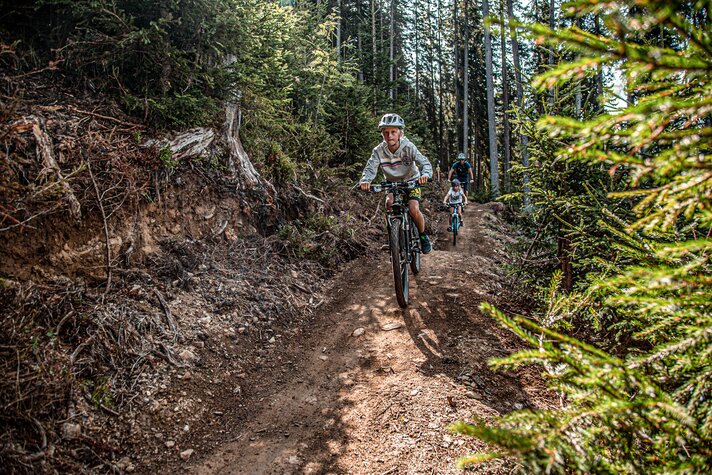 Eine Junge im Vordergrund und zwei Personen im Hintergund fahren durch den Wald im Bikepark am Hochjoch in der Silvretta Montafon.  | © Silvretta Montafon - Friederike Weber