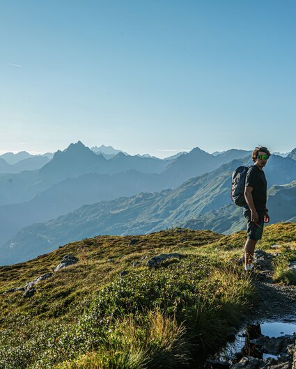 Ein Mann wandert in den Bergen im Sommer mit Aussicht auf die umliegenden Gipfel. | © Silvretta Montafon - Vanessa Strauch