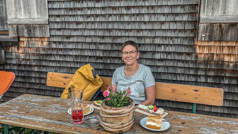 Eine Frau sitzt an einem rustikalen Tisch an einer Alpe mit Kuchen und Käsebrot. | © Silvretta Montafon - Vanessa Strauch