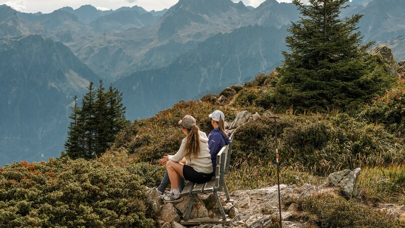 Zwei Frauen sitzen auf einer Bank auf einem Grat mit Weitblick im Herbst in der Silvretta Montafon. | © Silvretta Montafon - Vanessa Strauch
