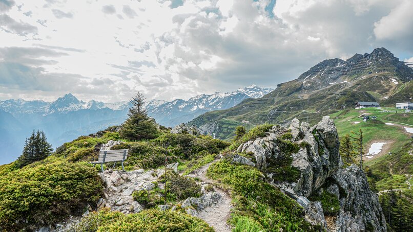 Ein Wanderweg an einem Grat mit Aussicht über die Berggipfel und einer Rastbank. | ©  Silvretta Montafon - Vanessa Strauch