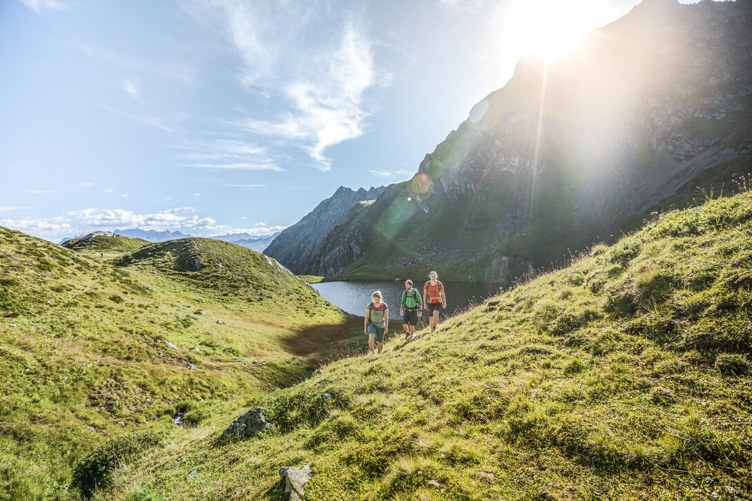 Drei Personen wandern bei Sonnenschein im Sommer in der Nähe eines Bergsees umringt von hohen Bergen. | © Silvretta Montafon - Stefan Kothner