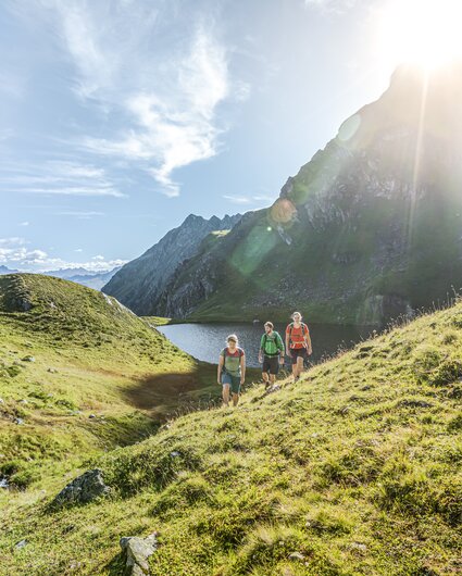Drei Personen wandern bei Sonnenschein im Sommer in der Nähe eines Bergsees umringt von hohen Bergen. | © Silvretta Montafon - Stefan Kothner