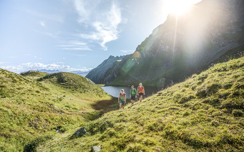 Drei Personen wandern bei Sonnenschein im Sommer in der Nähe eines Bergsees umringt von hohen Bergen. | © Silvretta Montafon - Stefan Kothner