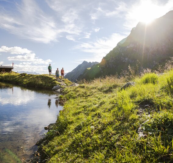 Drei Personen wandern bei Sonnenschein im Sommer in der Nähe eines Bergsees umringt von hohen Bergen. | © Silvretta Montafon - Stefan Kothner