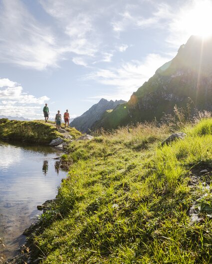 Drei Personen wandern bei Sonnenschein im Sommer in der Nähe eines Bergsees umringt von hohen Bergen. | © Silvretta Montafon - Stefan Kothner