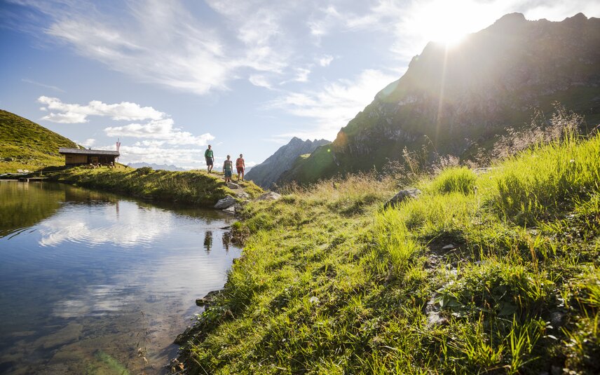 Drei Personen wandern bei Sonnenschein im Sommer in der Nähe eines Bergsees umringt von hohen Bergen. | © Silvretta Montafon - Stefan Kothner