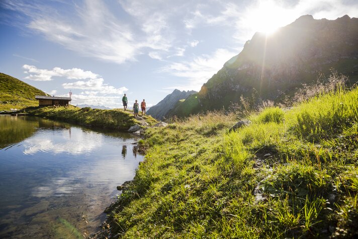 Drei Personen wandern bei Sonnenschein im Sommer in der Nähe eines Bergsees umringt von hohen Bergen. | © Silvretta Montafon - Stefan Kothner