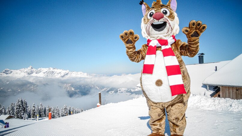 Das Maskottchen Monti Lux steht am Berg im Schnee und winkt in die Kamera. | © Silvretta Montafon