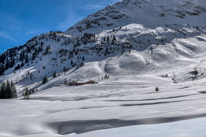 Verschneite Winterlandschaft in den Bergen mit einer Alp im Hintergrund. | © Silvretta Montafon - Vanessa Strauch