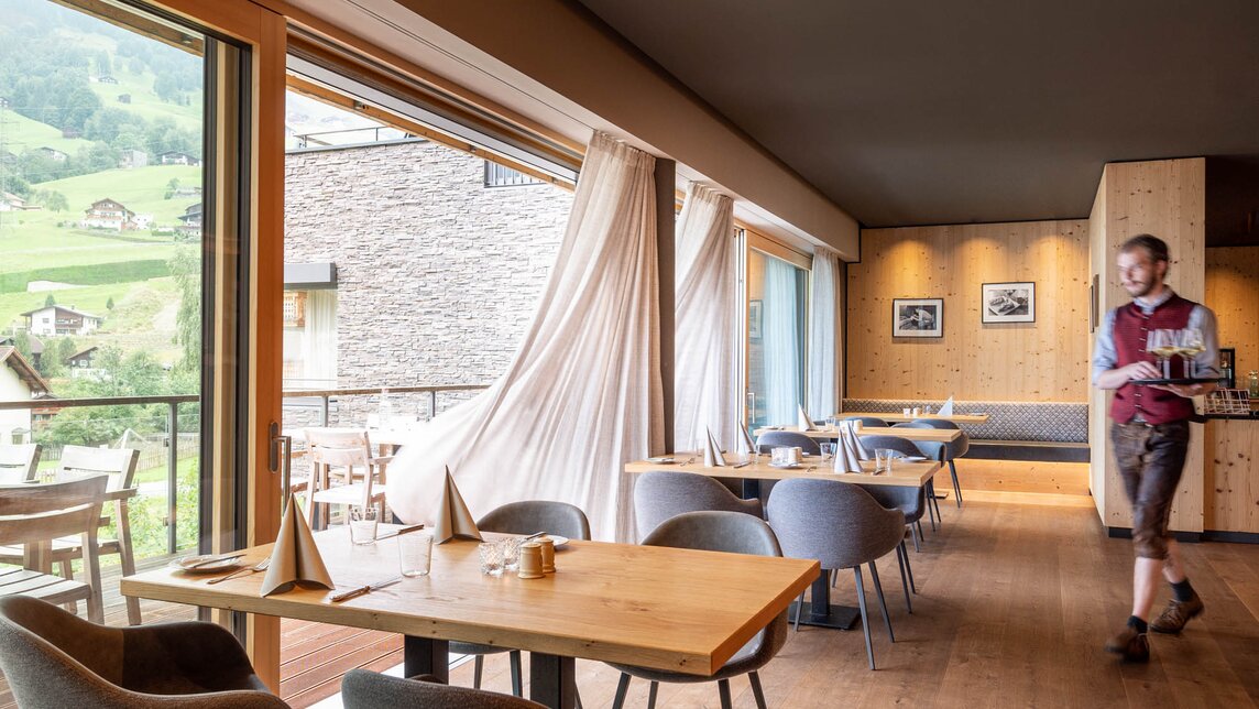 Ein Kellner läuft durch die gemütliche Gaststube mit gedeckten Tischen. | © Silvretta Montafon - blateral