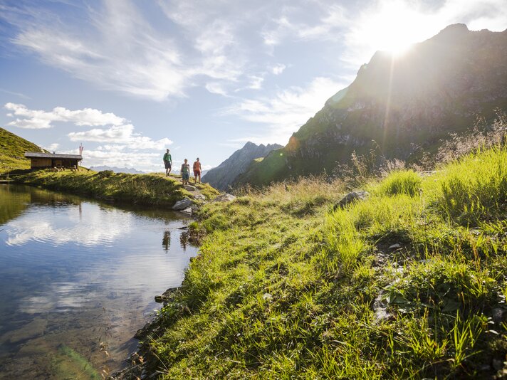 Drei Personen wandern bei Sonnenschein im Sommer in der Nähe eines Bergsees umringt von hohen Bergen. | © Silvretta Montafon - Stefan Kothner