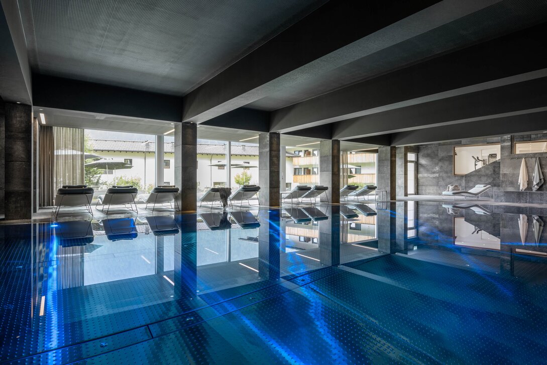 Der blaue Kristallpool im Inneren vom Sporthotel Silvretta Montafon mit Liegen und einer großen Fensterfront mit Blick ins Grüne | © Silvretta Montafon - blateral