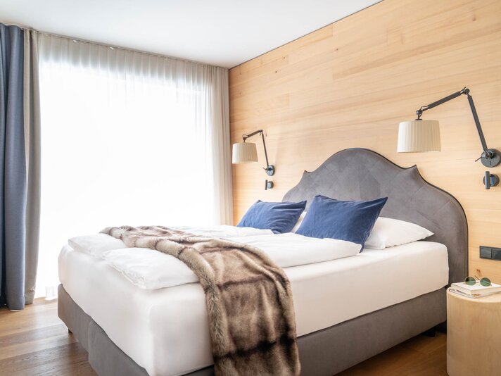 Ein großes Bett mit grauer Polsterung, einer weißen Decke und einem blauen Vorhang im Hintergrund | © Silvretta Montafon - blateral