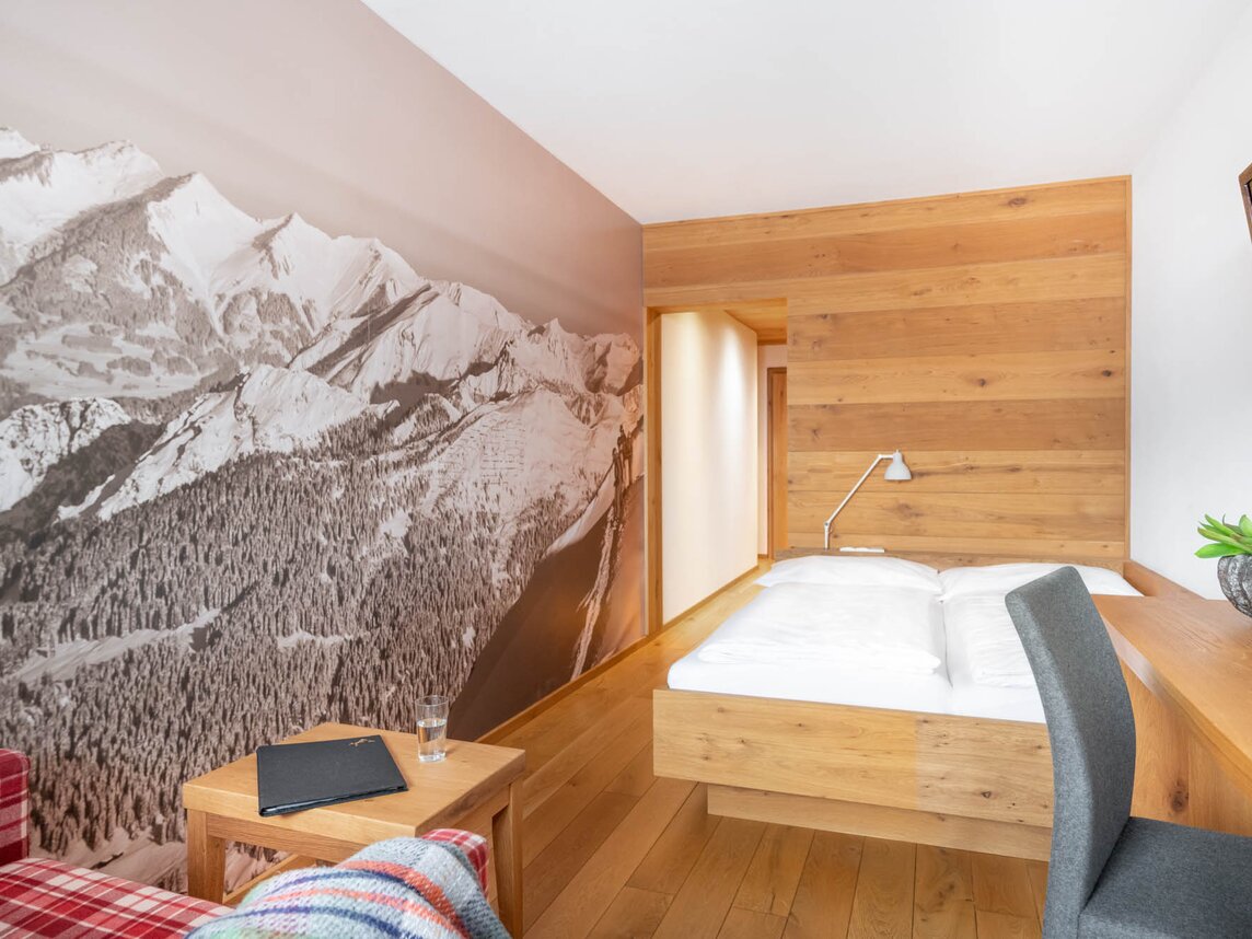 Ein Doppelbett mit Holzrahmen vor einer Holzwand und eine rot-karierter Couch | © Silvretta Montafon - blateral